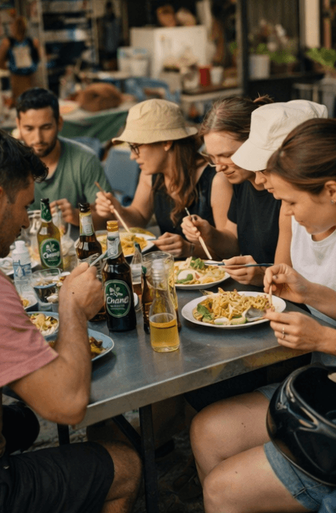 Friends enjoy Asian street food and beer at a casual outdoor table with chopsticks.