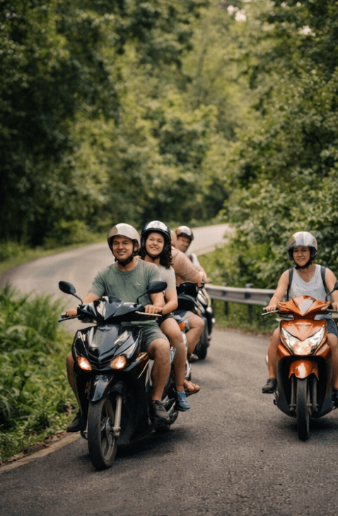 People wearing helmets ride scooters on a winding road through a lush, green forest.